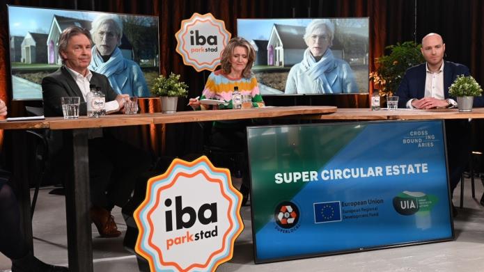 Het panel van de closing conference achter een tafel met op de voorgrond he IBA-logo en op de achtergrond burgemeester Dassen-Housen op een t.v.-scherm