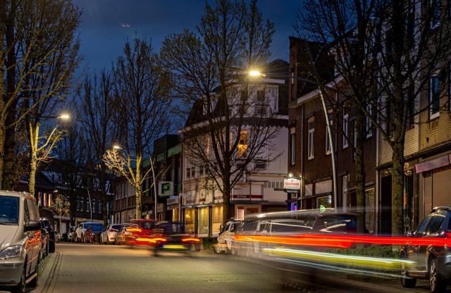 Twee straatlantaarns met LED verlichting  die branden in het donker in een straat met huizen, er komen auto's voorbij die met lange sluitertijd gefotografeerd zijn